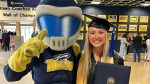 Business Gradate Student Molly Bennett, dressed in graduating cap and gown and holding a diploma, poses with UToledo mascot Rocky.